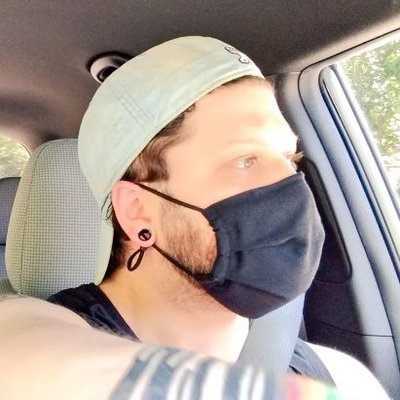 Picture of John wearing a UNC baseball cap backwards and a black cloth mask inside a car, looking away from the camera